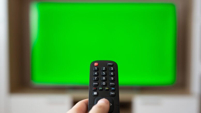 A hand with a TV remote control over a green screen. A place for your advertising